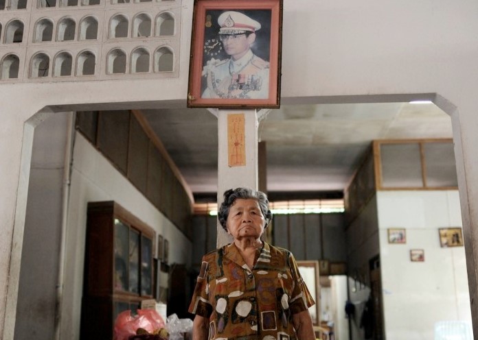 An elderly Asian widow stands below the photo of her deceased husband in his army dress uniform